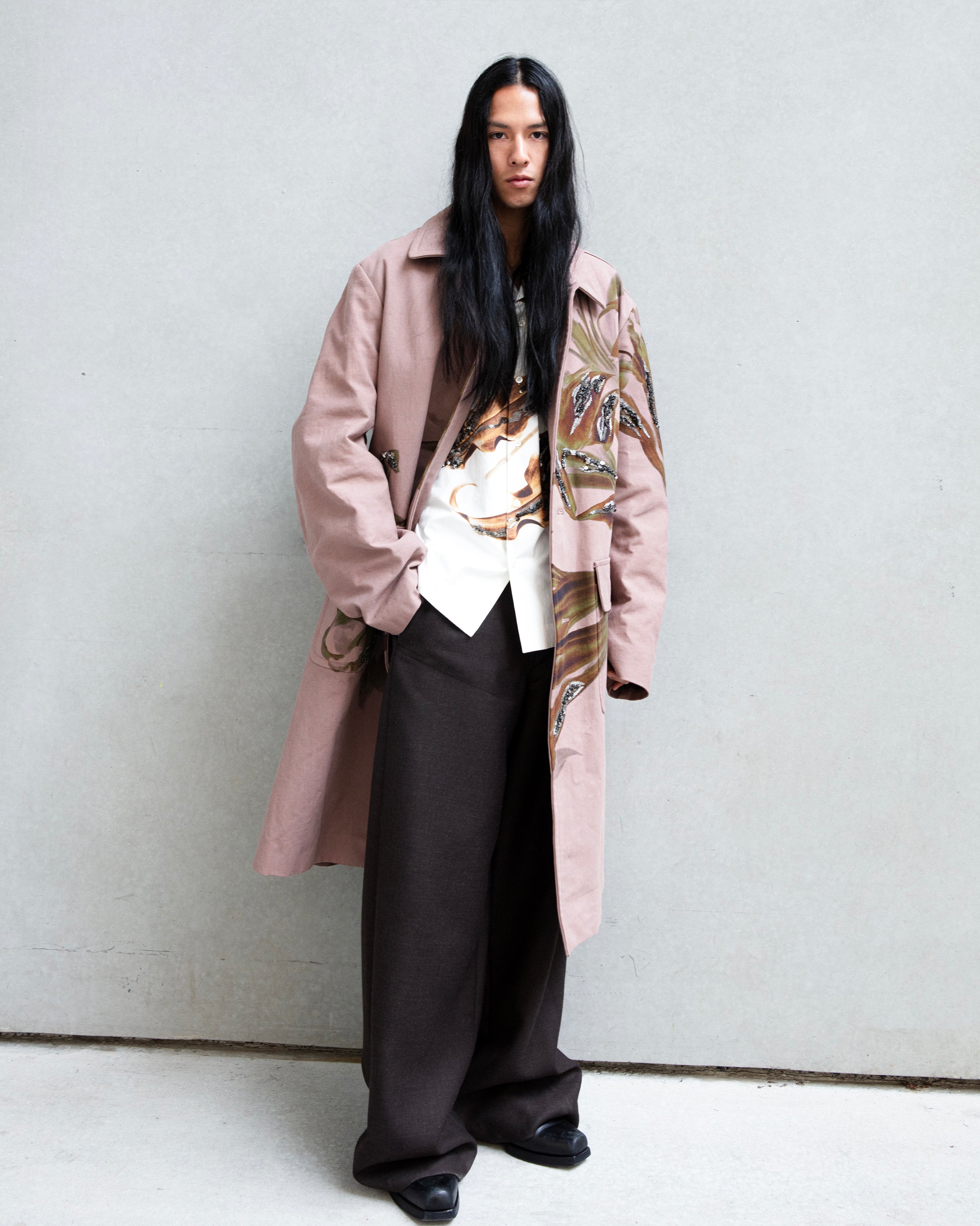 Male model wearing a blush pink Embellished embroidered coat over the Carlty Embellished printed shirt and Long Wool wide-leg trousers by Dries Van Noten, photographed against a minimalist gray wall for Atelier New York’s Men’s Collection.