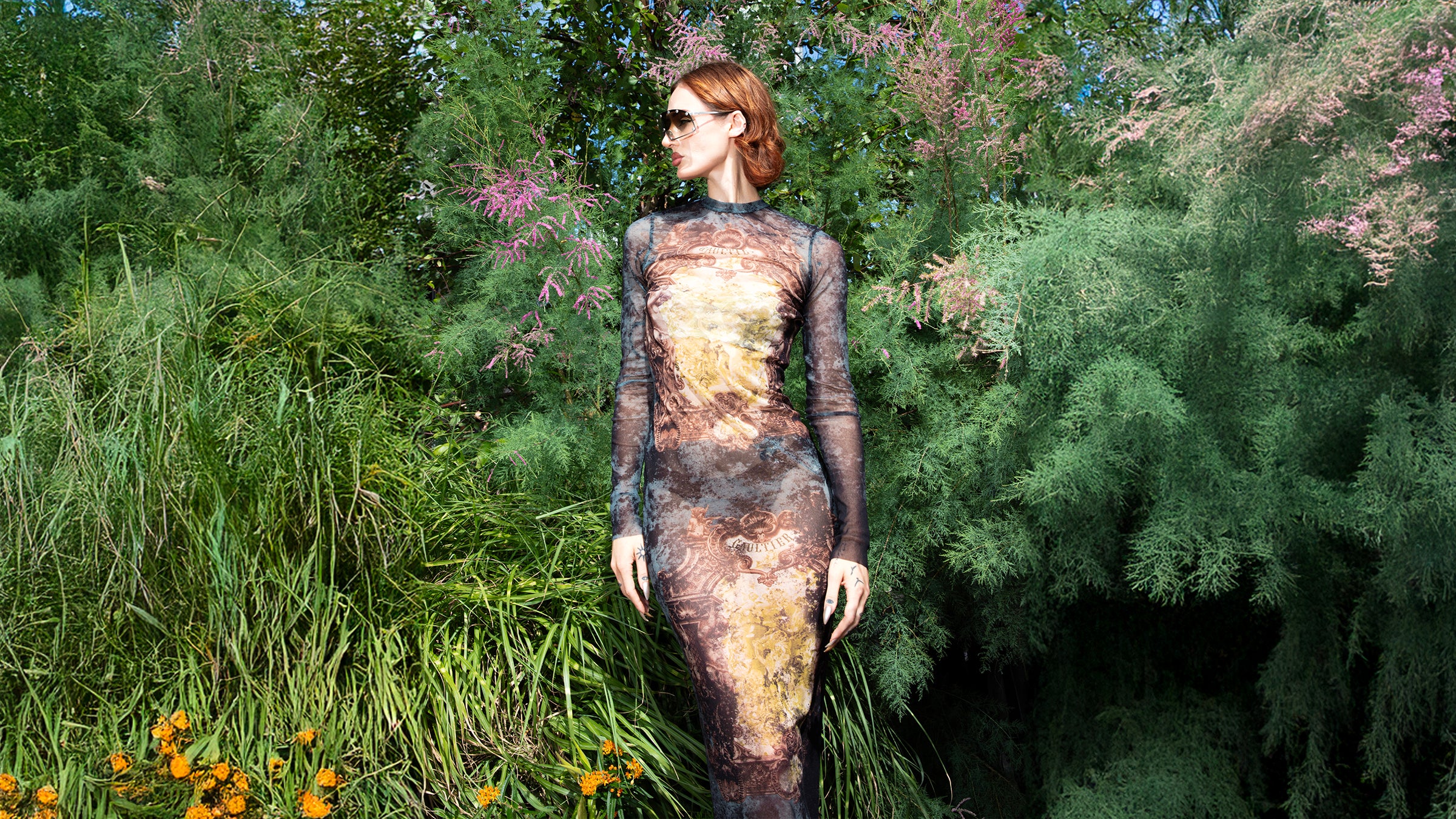 Female model wearing a baroque print sheer long-sleeve dress by Jean Paul Gaultier, standing in a lush green garden surrounded by tall grasses and pink flowering plants.